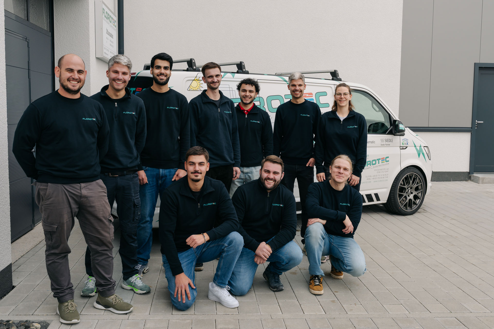 Eine Gruppe lächelnder junger Menschen steht und kniet vor einem weißen Firmenwagen