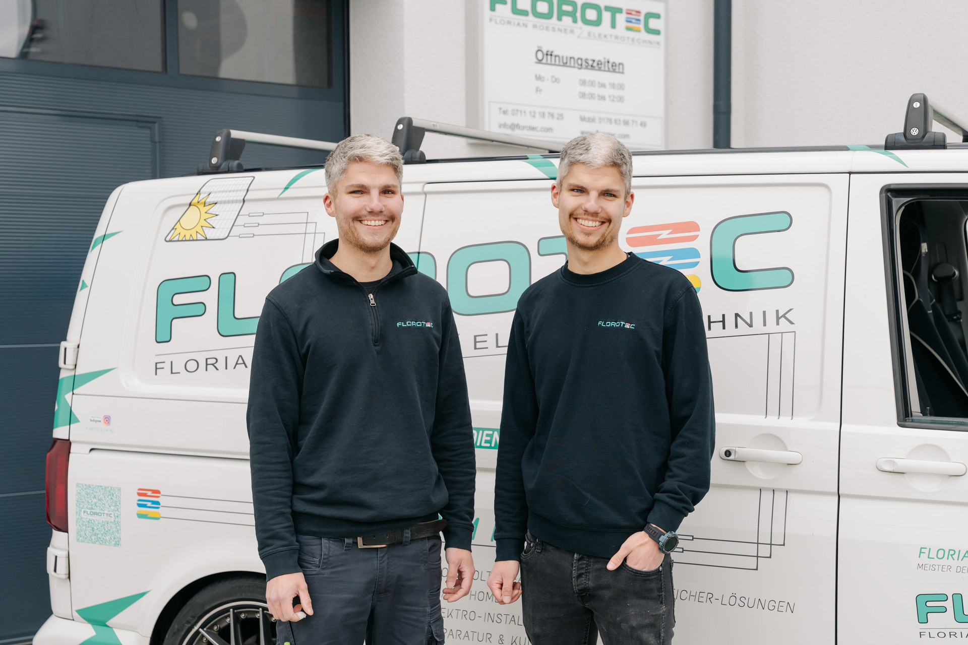 Zwei junge Männer mit kurzen grauen Haaren lächeln freundlich vor einem Firmenwagen