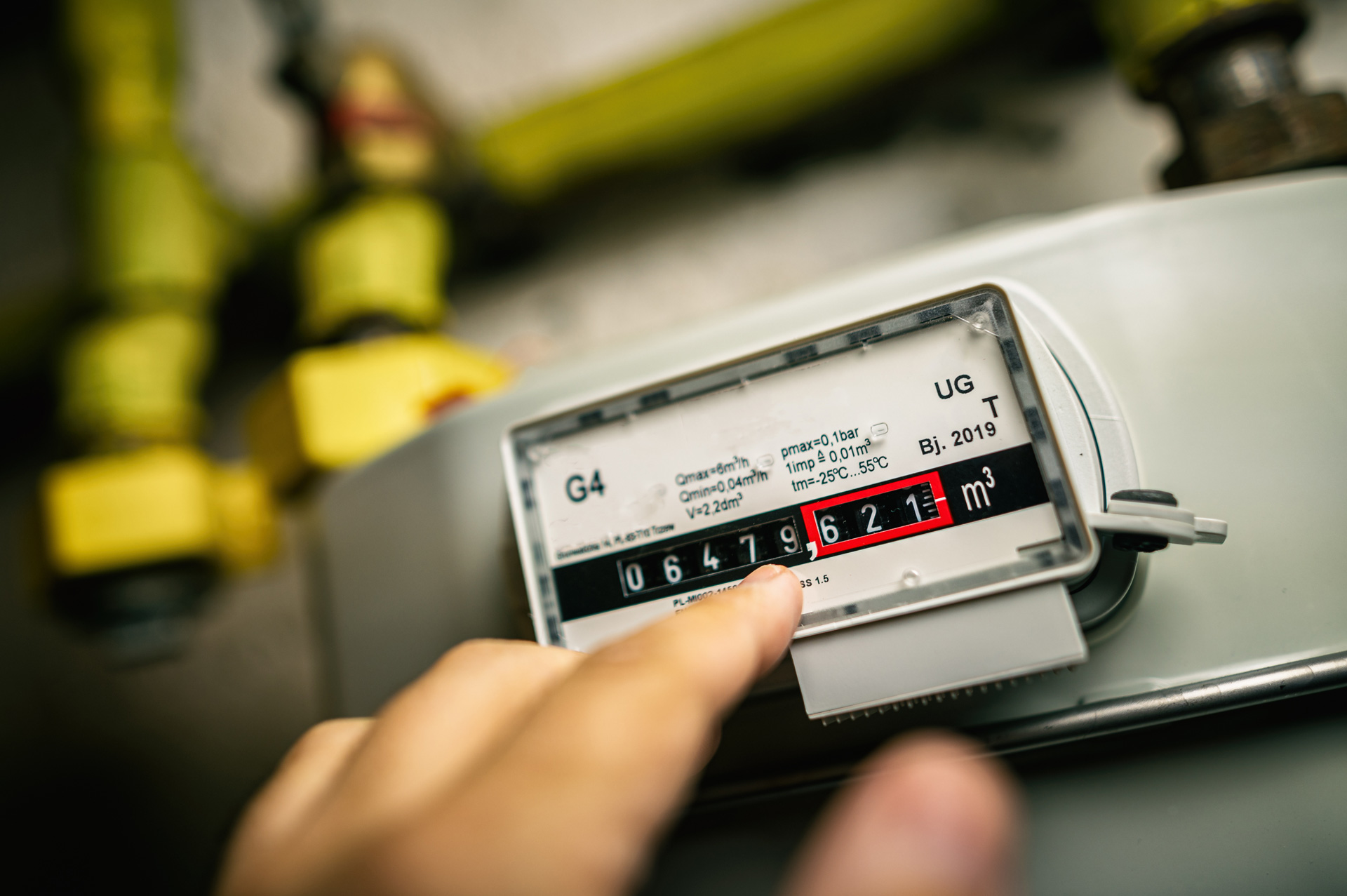 Eine Hand zeigt auf einen Gaszähler mit gelben Rohren und schwarzen Zahlen