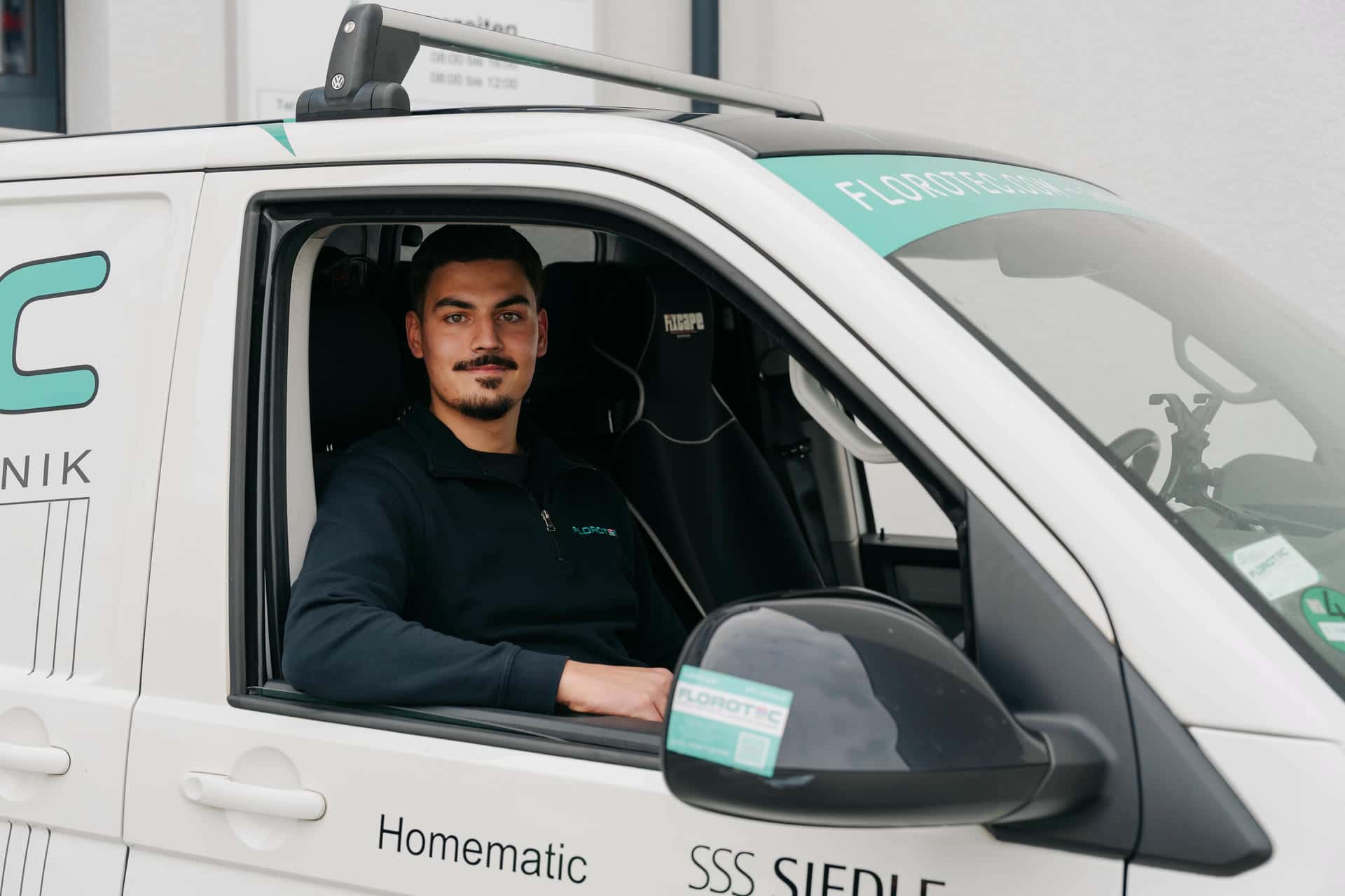 Junger Mann mit Bart in schwarzer Jacke sitzt in weiss tuerkisem Firmenwagen
